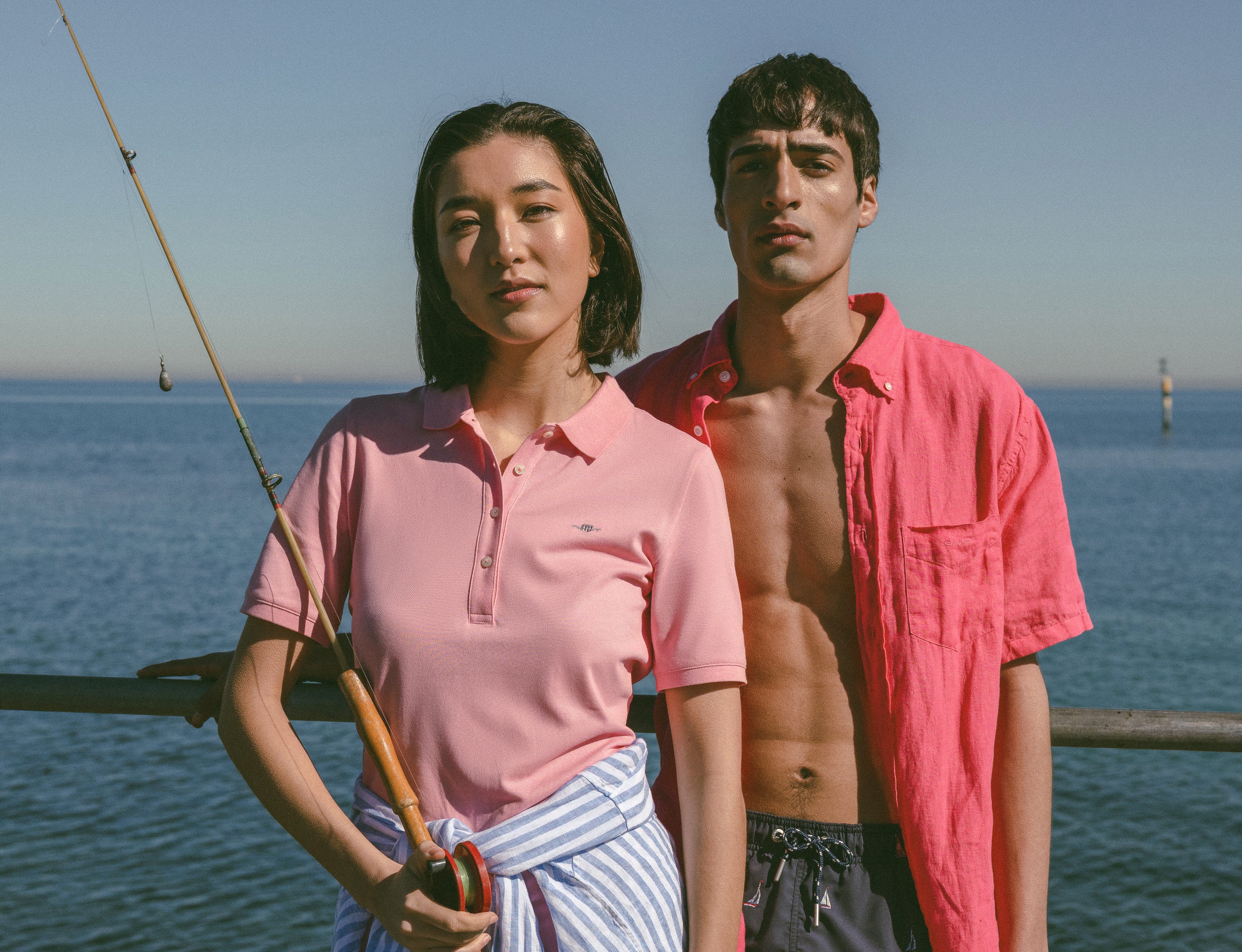Two people by the seaside, wearing a GANT polo dress and a GANT shirt with swim shorts. One is holding a fishing rod.