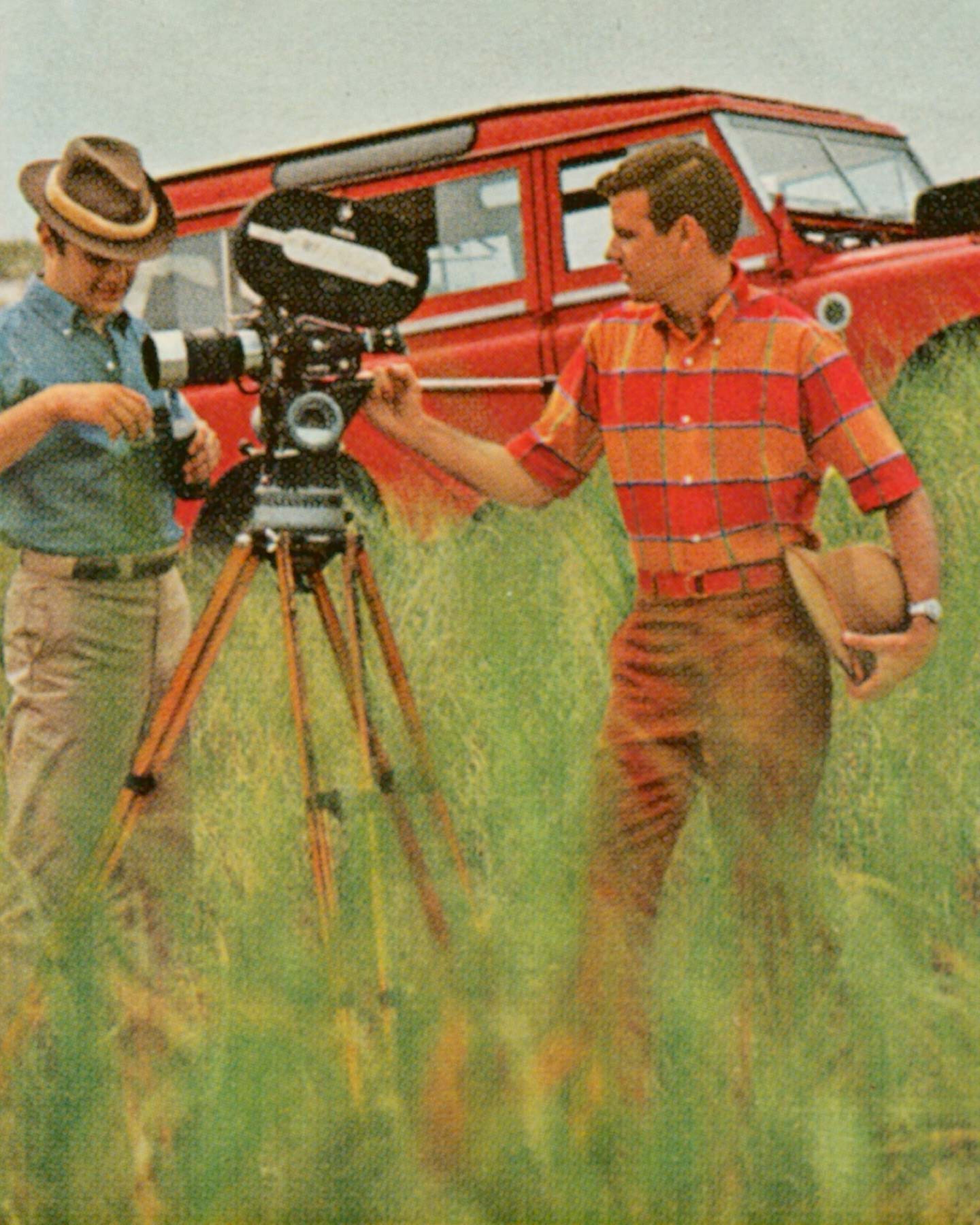 Two men with vintage film equipment in a field, with a red car in the background.
